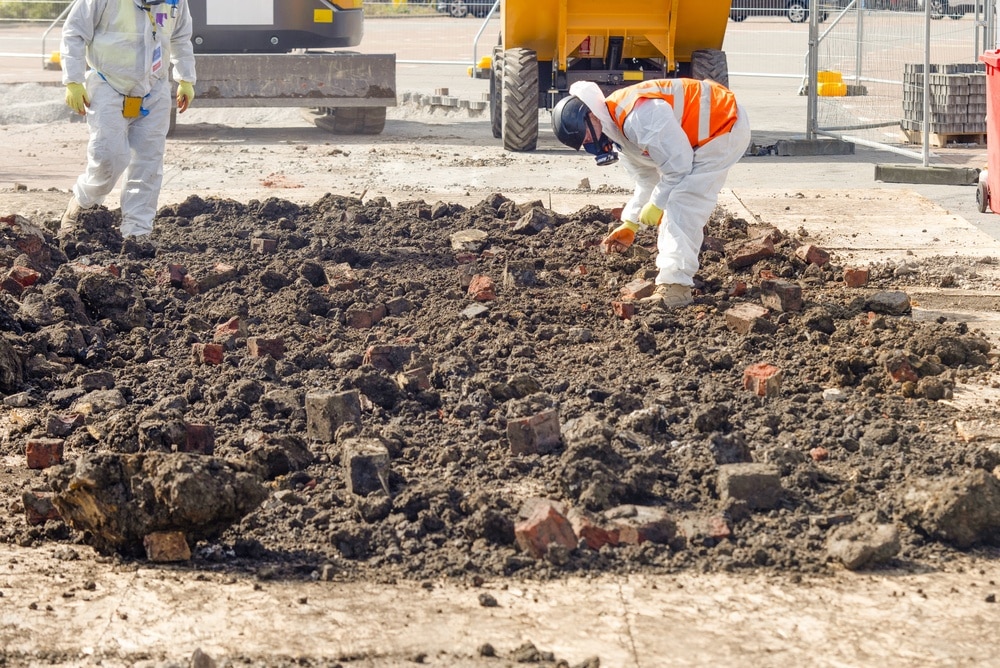 Two Workers In Protective Gear Remove Asbestos From Contaminated Ground